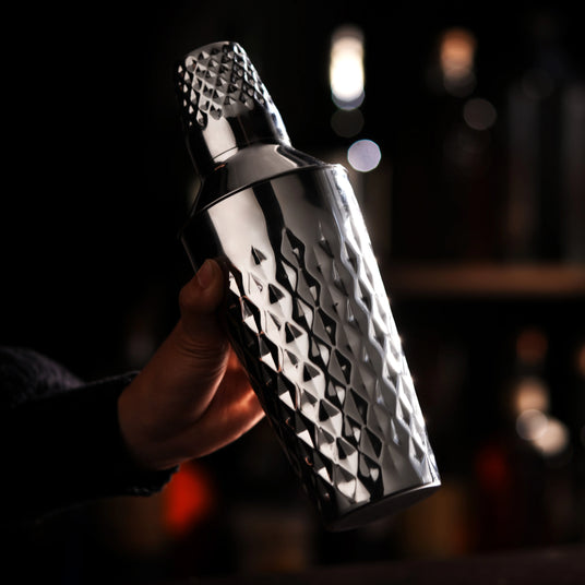 Metallic cocktail shaker held by a hand against a blurred bar background